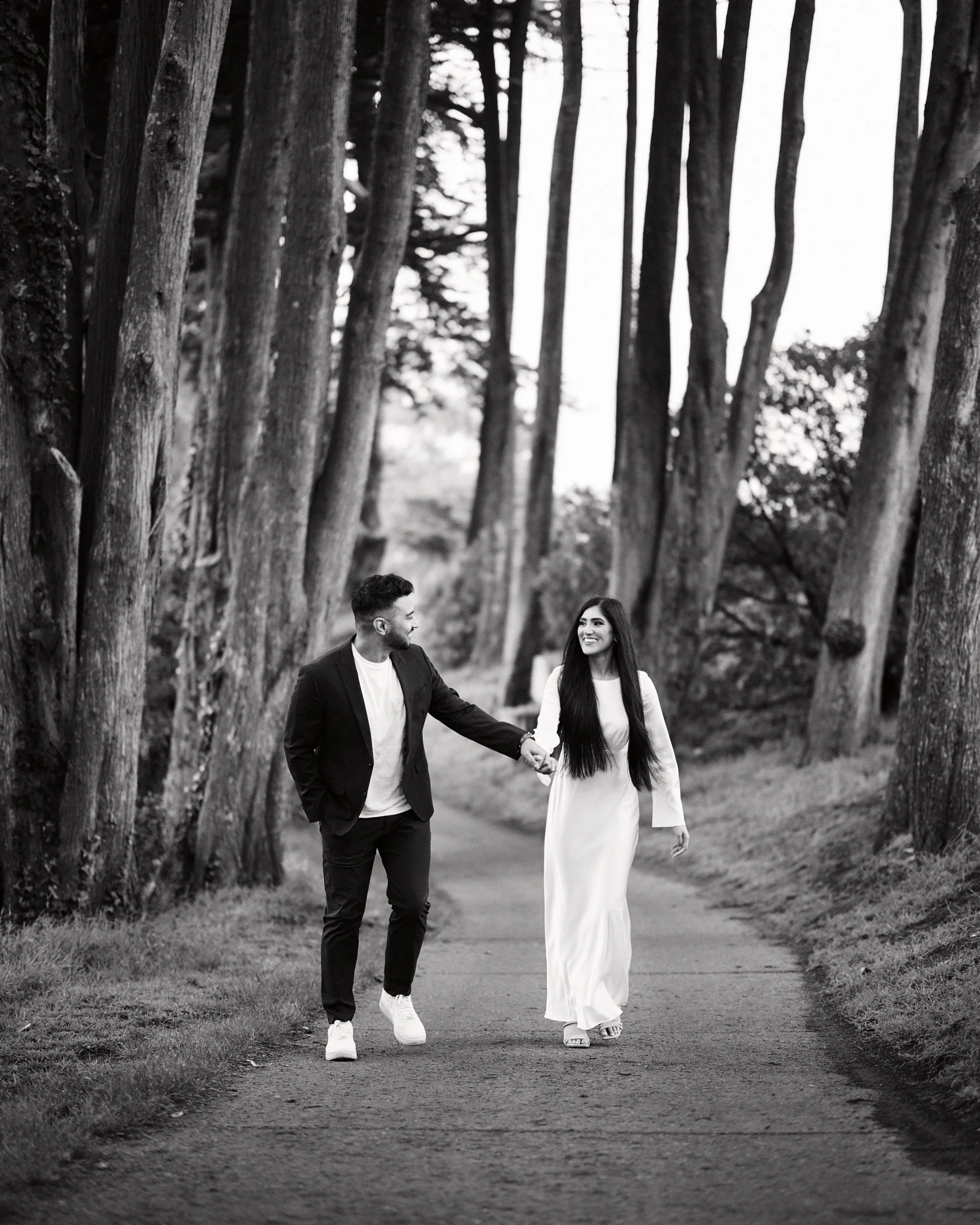 A couple holding hands on a path beneath tall cypress trees, in black and white